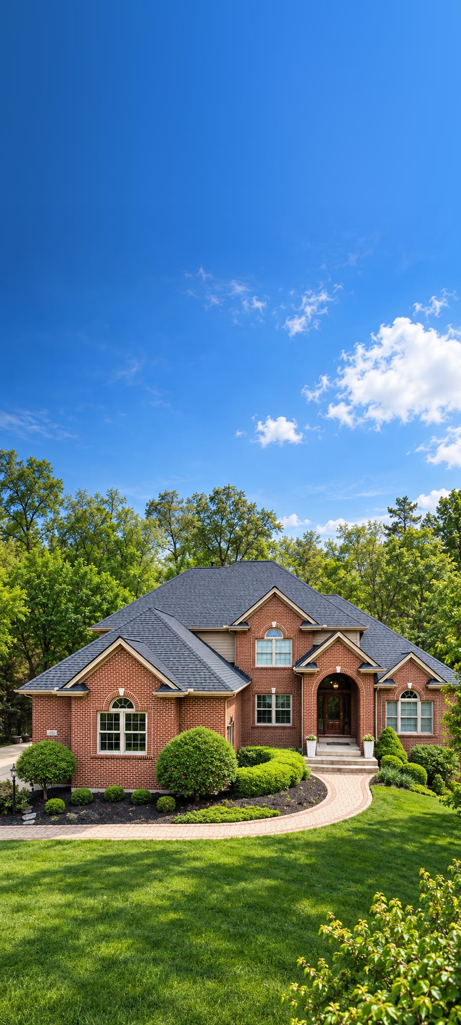 A brick estate under a blue sky with a charcoal architectural shingle roof, trimmed shrubs along the front and a paver walk to the door