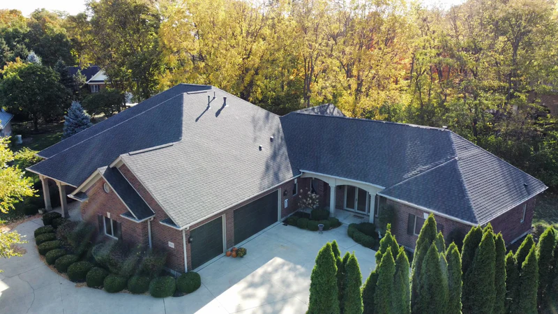 Drone view of a brick home with a new pewter shingle roof at the edge of the woods